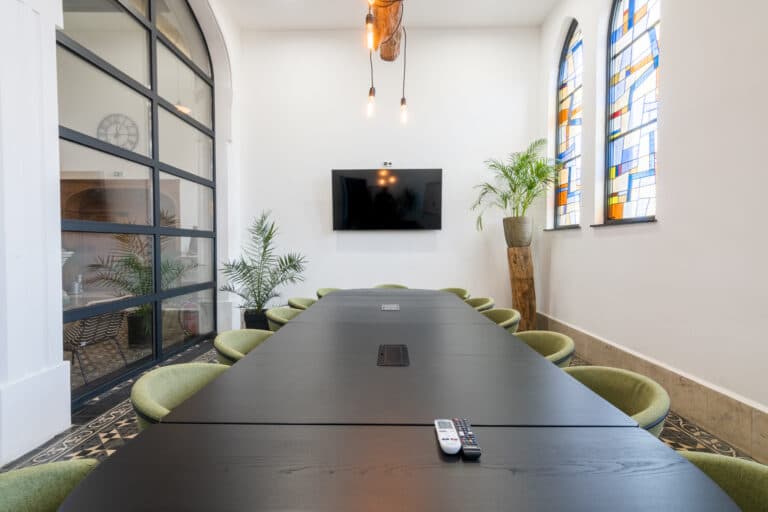 Salle de réunion moderne avec une grande table noire, des chaises vertes, un téléviseur et des fenêtres en vitrail.