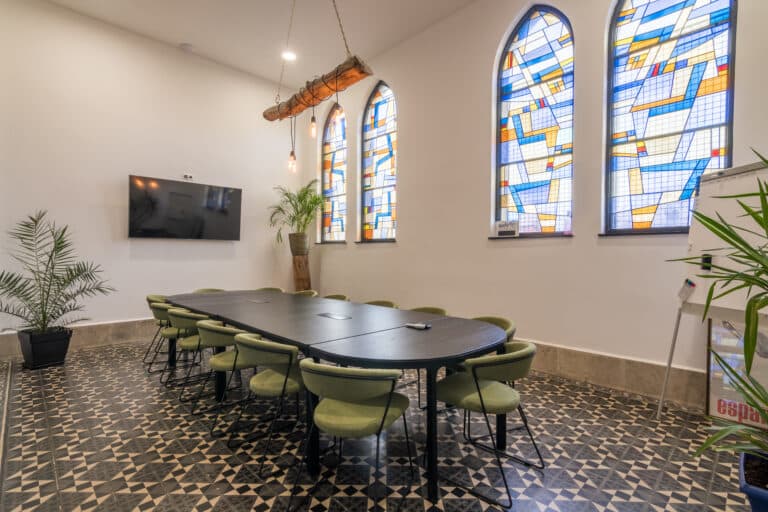 Salle de réunion moderne avec une grande table, des chaises vertes, un écran et des vitraux colorés.