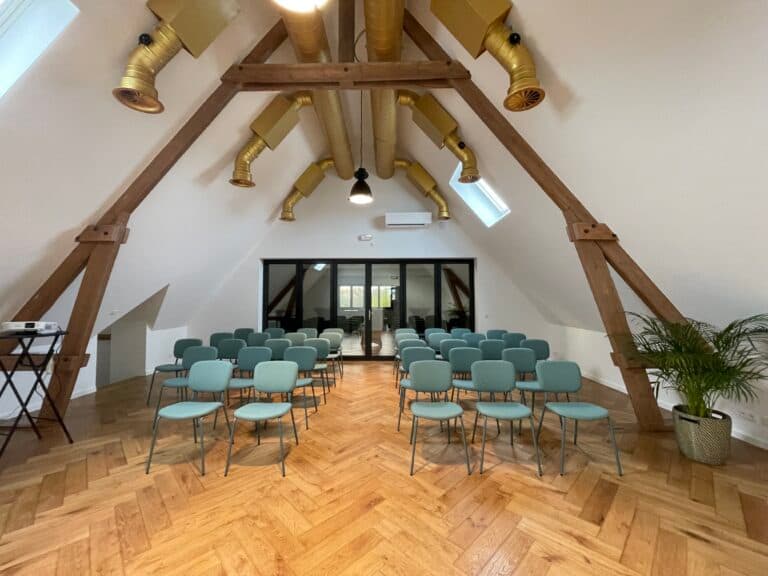 Salle de réunion avec des chaises vertes alignées et un éclairage dans un espace à la charpente en bois, avec des fenêtres et un parquet en bois.