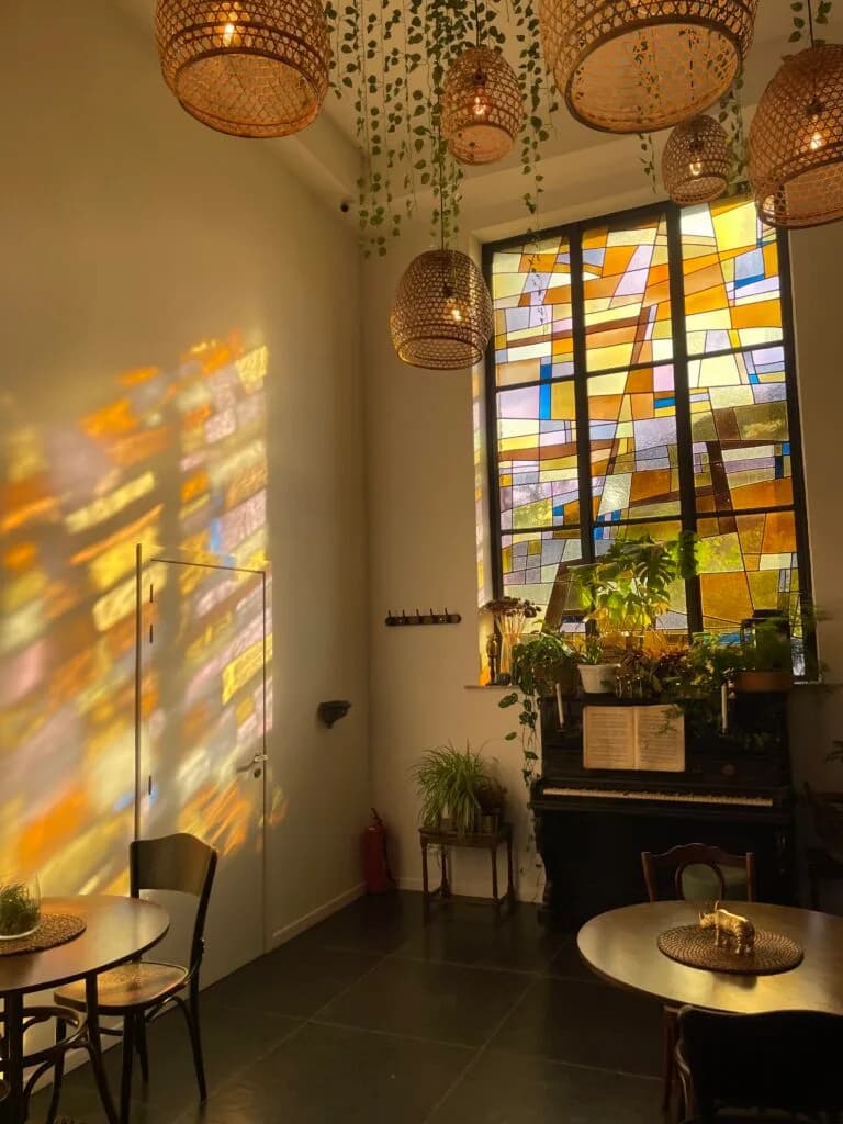 Un intérieur lumineux avec des tables, des plantes, un piano ancien et une grande fenêtre vitrée colorée projetant des reflets dans la pièce.