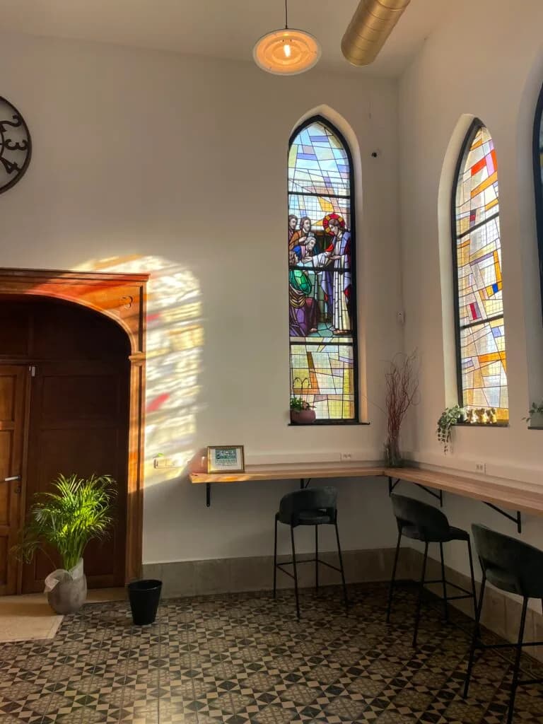 Intérieur d'un espace avec des murs blancs, des fenêtres en vitrail coloré, des chaises noires autour d'un bar, et une végétation décorative.